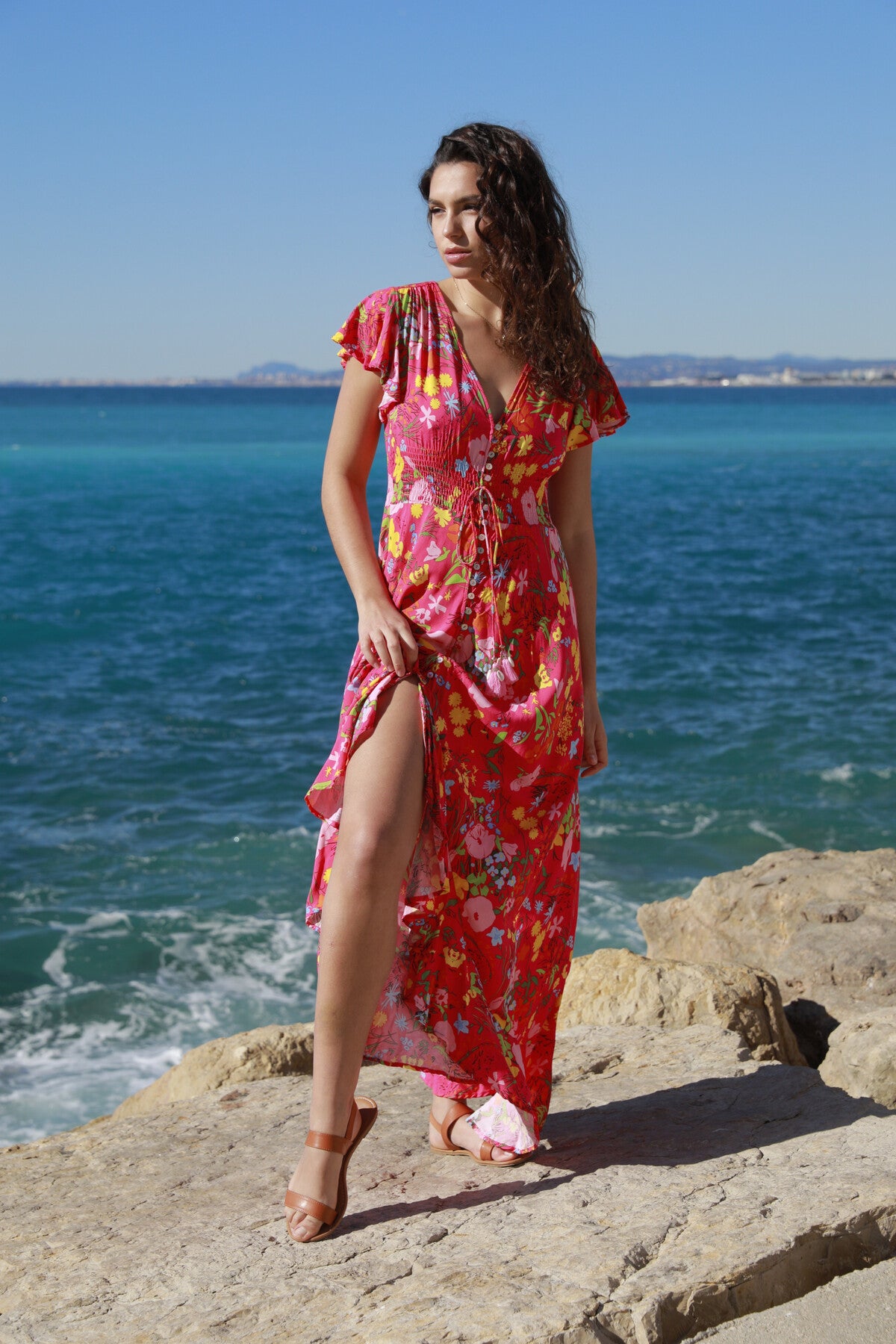 A woman wearing The Belle Boutiques Pink Flash Boho Flower Dress with ruffle detail stands on rocks by the sea, gazing left. Her wavy hair and sandals enhance the serene scene of a clear sky and calm ocean.