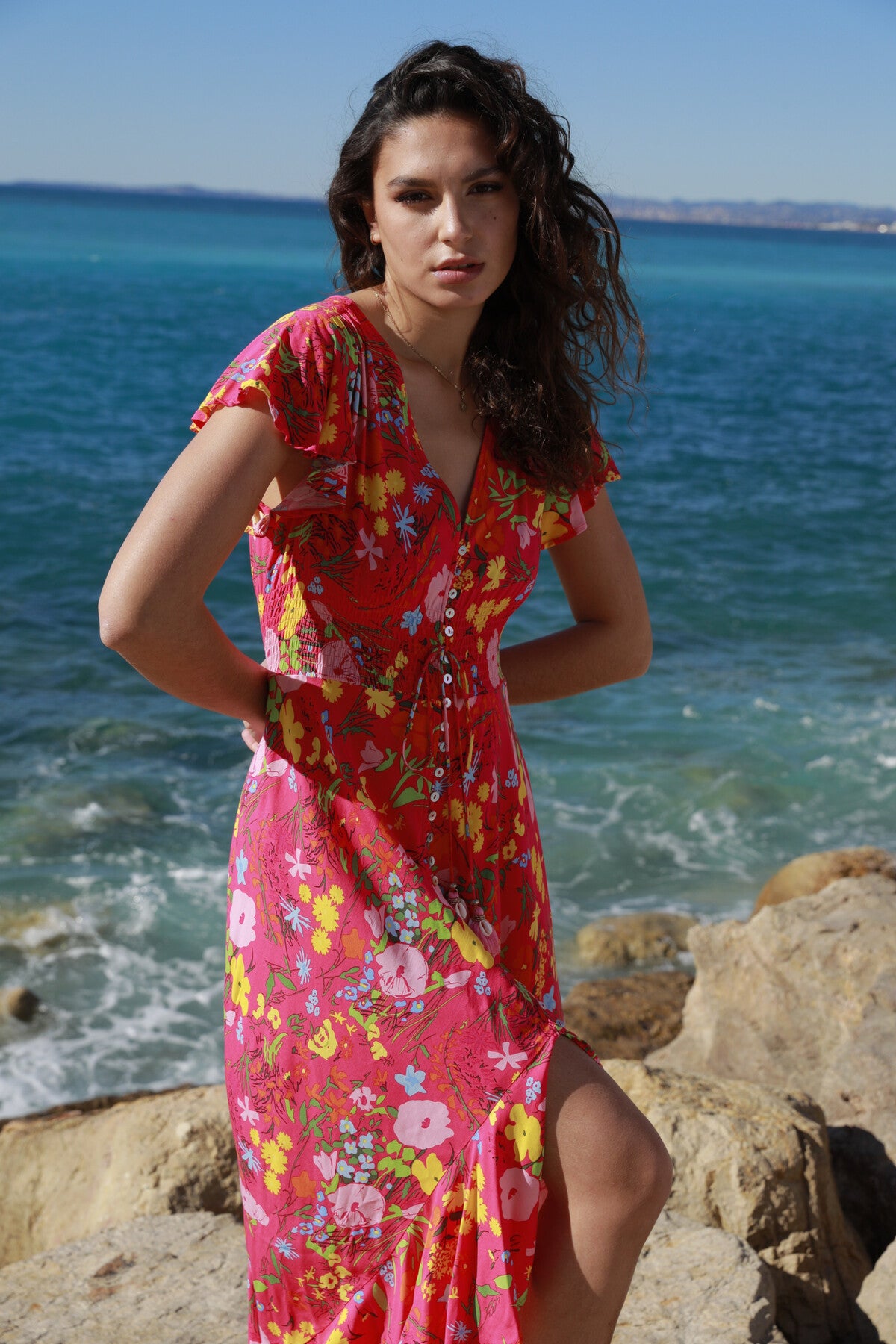 Standing on rocky terrain by the ocean, a woman exudes holiday elegance in The Belle Boutiques Pink Flash Boho Flower Dress. Her vibrant red dress with ruffle details complements the calm sea and clear blue sky. Her windy curls frame her face as she gazes toward the camera.