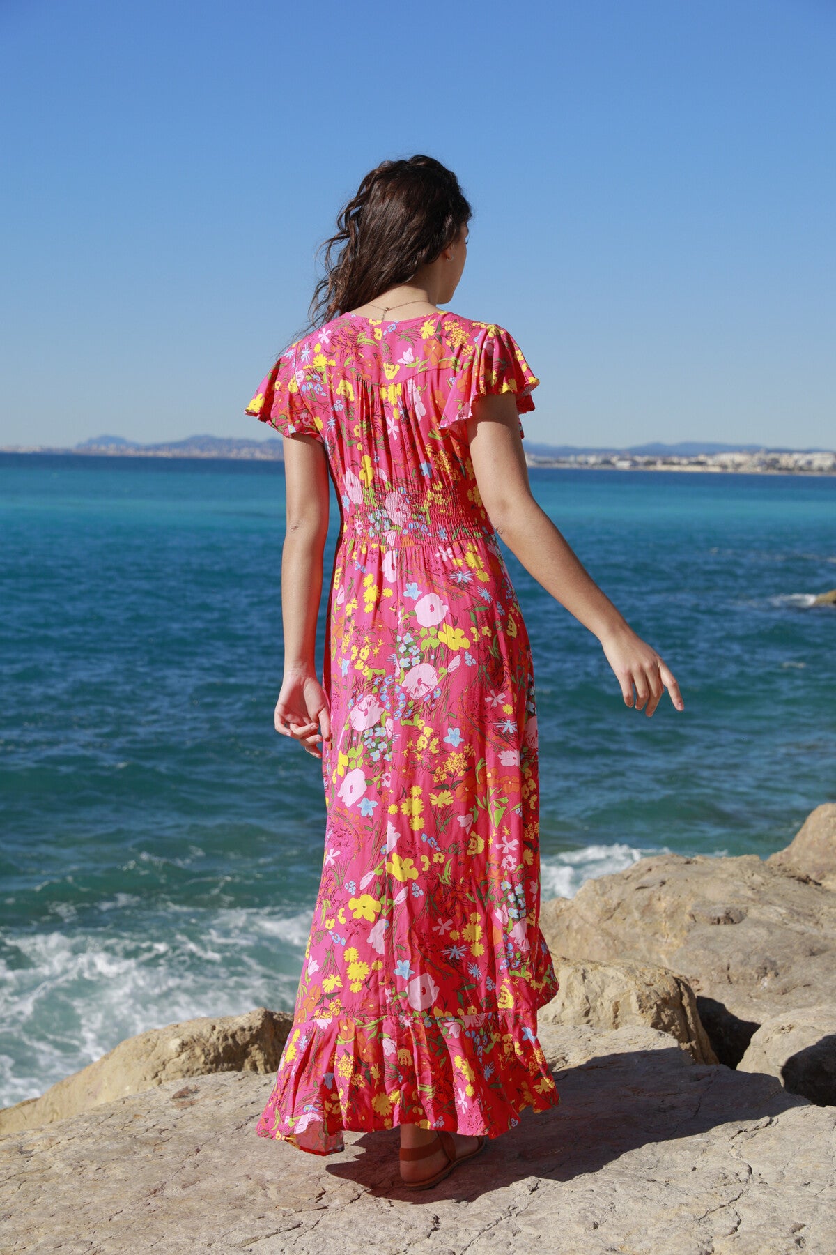 A woman with long brown hair stands on ocean rocks, wearing The Belle Boutiques Pink Flash Boho Flower Dress. Its ruffles dance in the breeze as she faces the sea under a clear blue sky, creating a serene holiday scene.