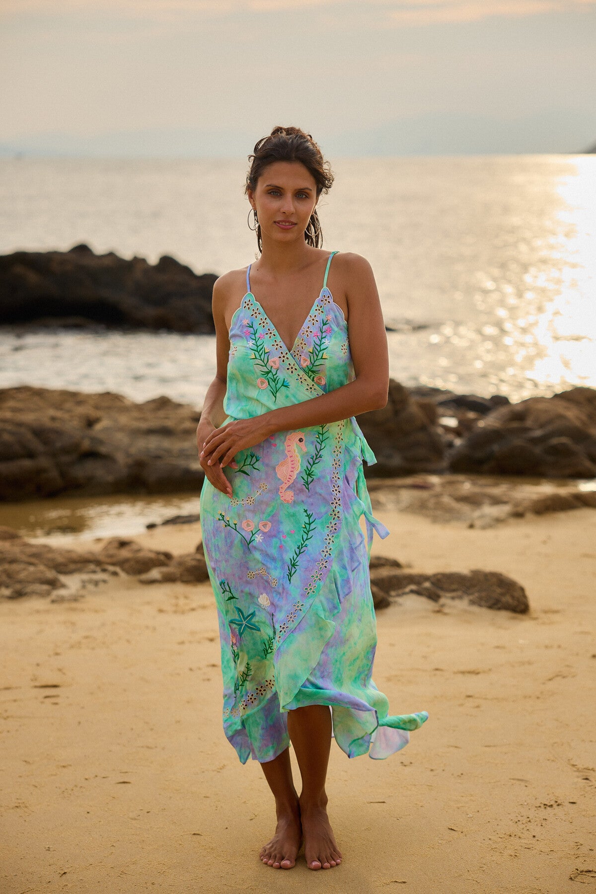 At sunset, a person stands barefoot on a sandy beach in a flowing, mid-length Tie Dye Wrap Dress by The Belle Boutique, showcasing divine green and blue hues. The ocean sparkles against rocky shores under a soft, hazy sky.
