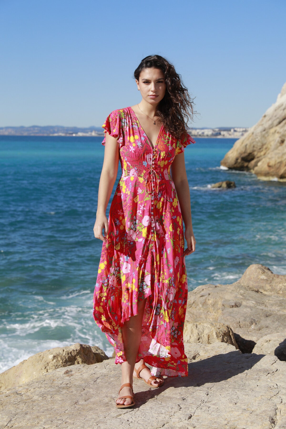 A woman stands on rocky terrain by the ocean wearing The Belle Boutiques Pink Flash Boho Flower Dress. Her vibrant holiday dress, adorned with multicolored patterns and ruffle detail, sways in the breeze. Behind her are blue waters and cliffs under a clear sky, as she wears sandals with flowing hair.