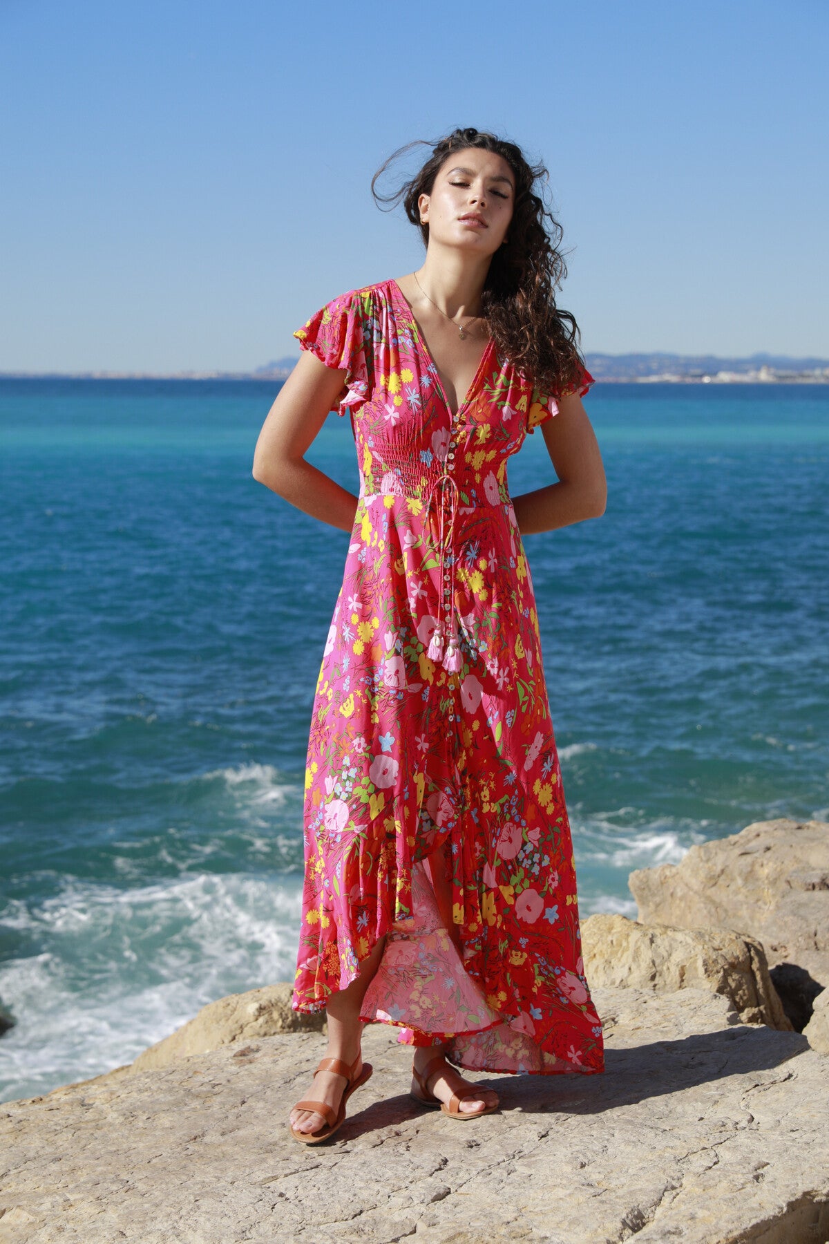 A woman in a Pink Flash Boho Flower Dress by The Belle Boutique stands on a rocky shore, the ruffle detail dancing in the ocean breeze against the clear blue sky.
